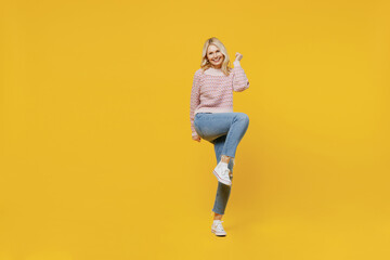 Fototapeta na wymiar Full body elderly fun excited blonde overjoyed woman 50s in pink sweater do winner gesture clench fist raise up leg isolated on plain yellow color background studio portrait. People lifestyle concep.