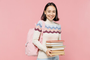 Smiling happy fun teen student girl of Asian ethnicity wearing sweater backpack hold pile of books look camera isolated on pastel plain light pink background Education in university college concept.