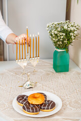 A female hand lights candles in Hanukkah on a table near donuts and a vase of flowers.