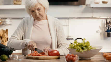 senior woman cutting fresh bell pepper near lettuce and cherry tomatoes in kitchen.