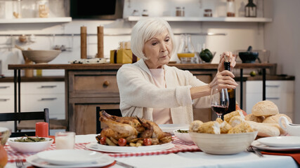 elderly woman uncorking bottle of red wine near roasted turkey, grilled corn and buns.