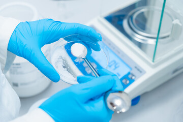 Digital analytical balance for accurate weighing of samples. Close up scientist hands in rubber gloves use metal spatula for weighing white powder of the substance.
