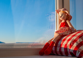 A doll in a red dress sits on the windowsill and looks out the window. Children loneliness concept....