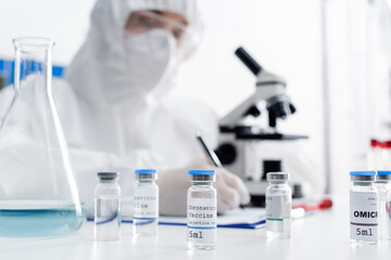 Fototapeta premium selective focus of vials with coronavirus vaccine near scientist writing near microscope on blurred background.