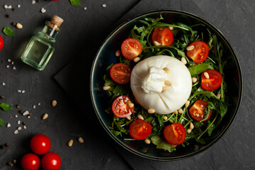 Salad with traditional italian burrata cheese with arugula and tomatoes om dark concrete table with cooking ingredients cherry tomatoes, basil, garlic and olive oil. Top view.