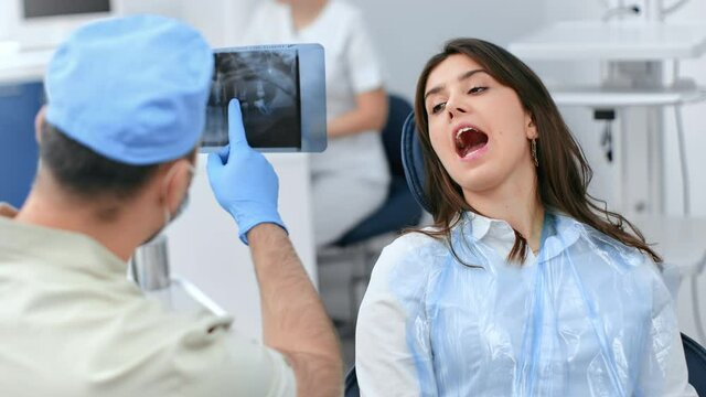 Male dentist pointing on x ray image consulting female patient open mouth on chair at stomatology clinic