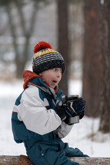 Fototapeta na wymiar Boy child makes funny faces sitting on a log in snowy park with thermal mug. Picnic in winter.