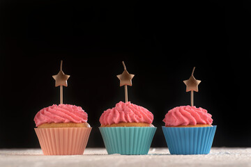 Three cupcakes with pink buttercream frosting with candles in the shape of star on black background. Copy space.
