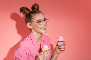 Funny smiling woman in pink shirt holding muffins on pink background. Sweets candy mood. Hard light. Copy space