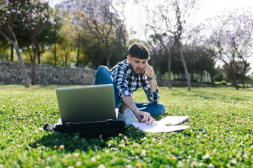 Young man sitting on the lawn studying on his laptop computer and talking on his phone