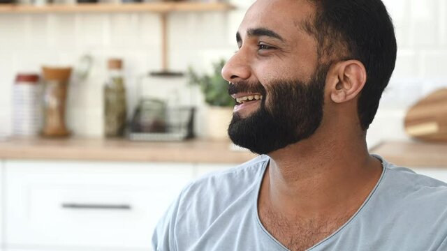 Healthy lifestyle. Positive healthy attractive Indian bearded man, stands at kitchen, drinks clean water from a glass, looks to the side, cares their health, drinks a daily norm of water, smiling