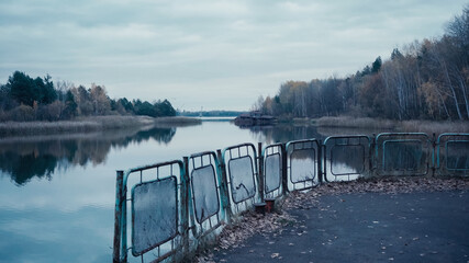 pripyat river and embankment with rusty fence under grey cloudy sky in chernobyl exclusion zone.
