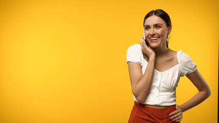 Smiling woman in blouse holding hand on hip and talking on smartphone isolated on yellow.