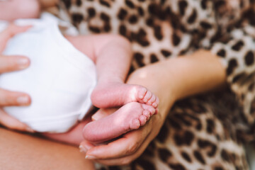 Beautiful mother in leopard robe breastfeed her newborn baby in white bodysuit holding feet in hand. Selective focus. 