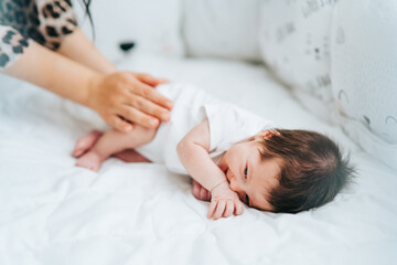 Cute little newborn baby with dark hair in white bodysuit lie sidelong smiling and mother touching his legs. Copy space.
