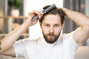 Close-up of a young positive caucasian guy in a white t-shirt combing his hair with a comb and looking at the camera. The concept of healthy hair, dandruff shampoo, hygiene and health