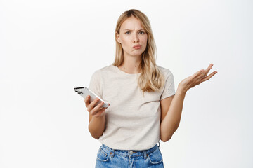 Confused blond woman shrugging with mobile phone, cant understand smth on smartphone, standing puzzled over white background - obrazy, fototapety, plakaty