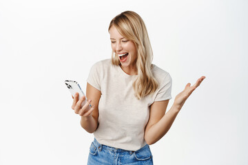 Happy blond girl winning on mobile phone app, watching smartphone and reacting with joy and amazement to notification, standing over white background