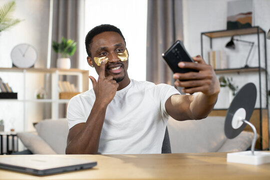 Close-up of a young positive African guy in a white t-shirt uses patches under his eyes pointing finger on the patch while making selfie on smartphone. The concept of healthy skin, hygiene and health