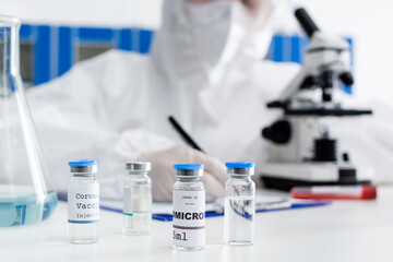 partial view of blurred doctor writing on clipboard near microscope and vials with covid-19 omicron...