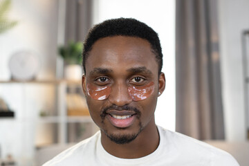 Front close up view of a positive handsome man. Cheerful stylish African guy in white T-shirt with pink collagen patches under the eyes smiling looking into camera