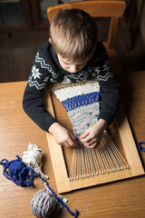 A boy weaves a rug from old T-shirts on a hand loom. Top view, vertical frame