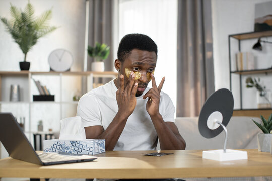 Stylish bearded athletic African American man puts gold patches under his eyes looking in the mirror. The concept of spending free time at home, men's health and beauty