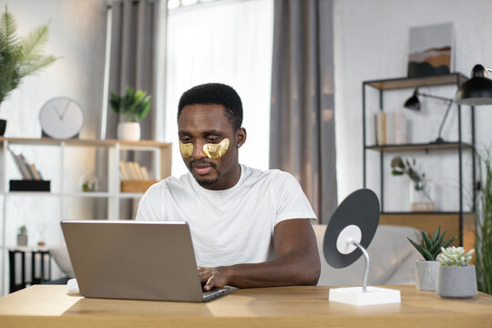 A happy successful smiling African man working at home on laptop sitting at a wooden table. A young man performs cosmetic procedures using gold patches while typing on a laptop