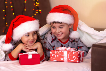 Two cute children, a girl and a boy, in pajamas and red hats, cuddle on white bed