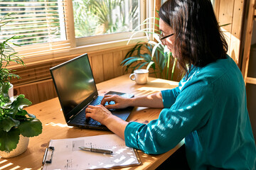 Serious brunette woman wearing eyeglasses sitting by window with hard sunlight in living room...