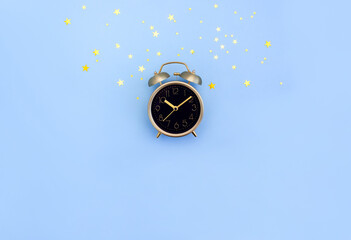 Golden retro alarm clock with black dial on blue background decorated with gold star confetti....