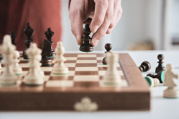 Chess game, hand moves black bishop across the chessboard. Male businessman plays chess in office. Business strategy, tactics concept. Wooden chess board, pawns, knights, rooks, bishops, queen, king
