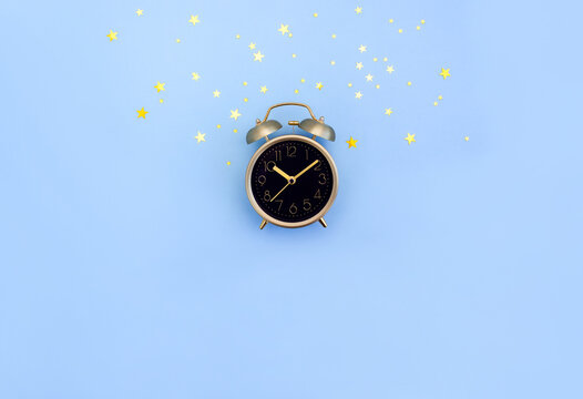 Golden retro alarm clock with black dial on blue background decorated with gold star confetti. Human circadian rhythms concept. Selective focus, copy space