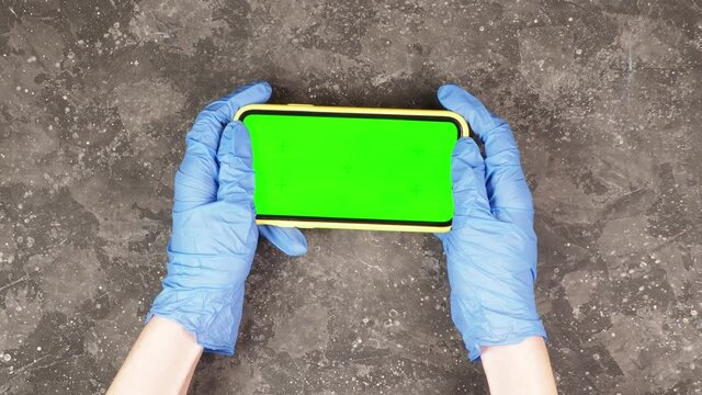 Hands of a young female doctor in blue protective gauntlet gloves holding a smartphone with a green screen mock up for an application on a gray background