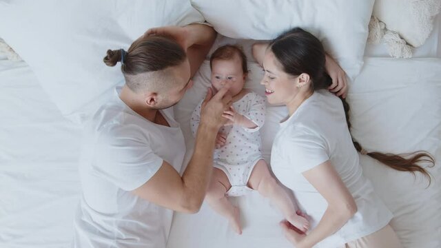 Loving and joyful young smiling caucasian parents, mother and father lying with small newborn baby on white bed sheet, gently play, kiss and touch nose. Happy family relationships concept. Top view