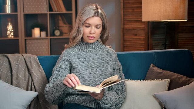 Happy domestic blonde woman in comfortable sweater reading paper book sitting on couch with torchere