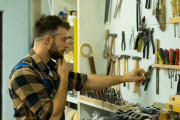 Young bearded caucasian carpenter holding hand on chin and thinking what tool to use for the work...
