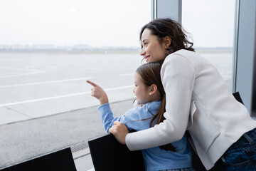 happy kid pointing with finger at window near cheerful mother in airport.