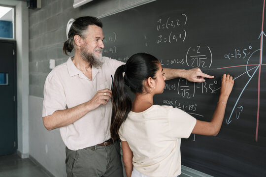 Maths Lesson In A Secondary School. Latin Female Teenager Student Writing On Blackboard With Help Of Friendly Teacher In High School. 