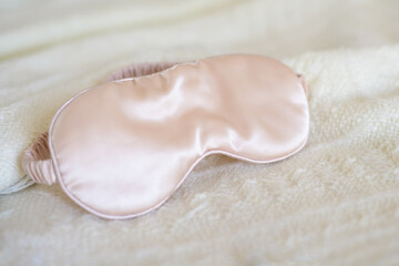 Apricot pink satin eye sleeping mask on beige knitted background. Insomnia and sleeplessness disorder concept. Close up, copy space, flat lay. Accessories for girls and women