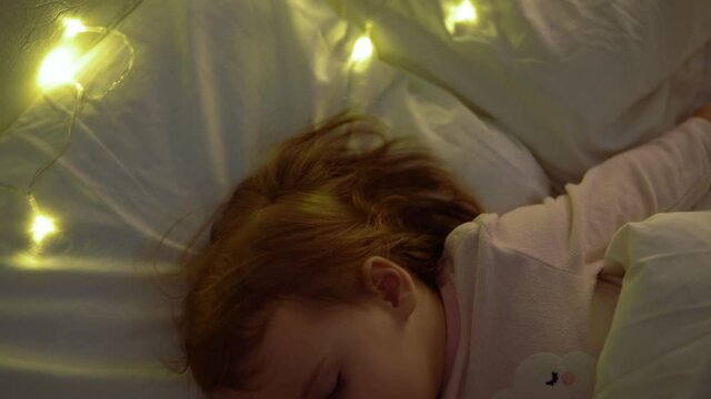 Cute Chuby Little Toddler Baby Girl 1-2 Years Old Sleeping In Pink Pajama On White Bed In Dark Bedroom. Child Have Relax Rest Time At Night Garland on Pillow. Childhood, Christmas, Parenthhood Concept