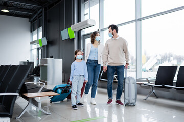 family in medical masks walking with baggage in airport lounge.