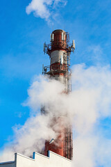 Industrial brick chimney with telecommunication equipment and white steam on blue sky
