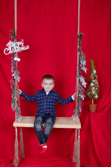 Fototapeta premium pretty blonde boy sitting on a wooden swing decorated with tree branches and christmas decor. on red background. new year theme.