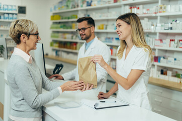Two pharmacist giving prescription medications to senior female customer in a pharmacy - obrazy, fototapety, plakaty