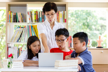 A Group of Asian Student Kid Reading and looking Laptop on the table with female teacher in School library with Shelf of Books  Background