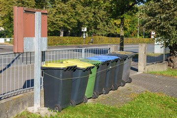 A group of plastic trash and recycle bins by a metal fence.