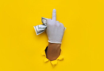 Fuck you gesture. A young doctor holds money in white medical gloves. Isolated. Yellow torn hole paper background.The concept of treatment fees, bribes, illegal surgery. Copy space.