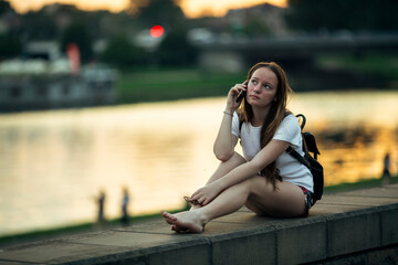A girl is anxiously talking on her phone while sitting on the city's waterfront at sunset.