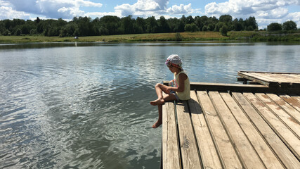 funny country alone barefoot child in cap sits on the edge of a wooden bridge over the water and...